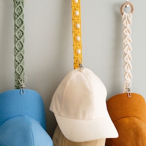 May include: Macrame hat organizers in green, yellow, and cream, each with a wooden ring and metal clip. Baseball caps in various colors, including blue, beige, and brown, are clipped to the organizers.