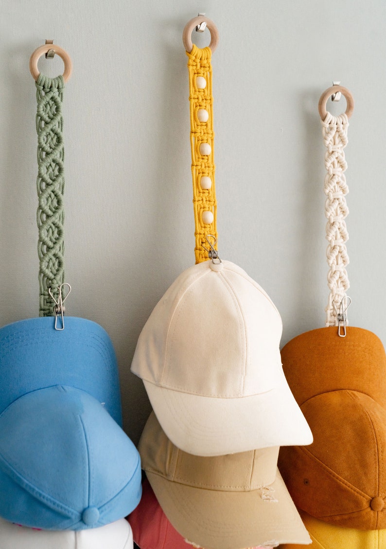 May include: Macrame hat organizers in green, yellow, and cream, each with a wooden ring and metal clip. Baseball caps in various colors, including blue, beige, and brown, are clipped to the organizers.