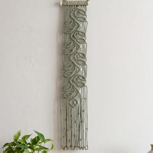 May include: A sage green macrame wall hanging featuring a series of interconnected leaf-shaped knots suspended from a horizontal wooden dowel. The design includes long vertical cords ending in small knots at the bottom edge of the piece.