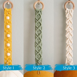 May include: Three macrame hat hangers in yellow, green, and cream colors. Each hanger has a wooden ring at the top and a metal clip at the bottom. Style 1 is yellow with wooden beads. Style 2 is green with a diamond pattern. Style 3 is cream.
