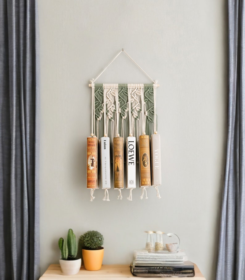 K&ouml;nnte beinhalten: Ein Wandbehang aus Makramee mit einem gr&uuml;n-wei&szlig;en Muster h&auml;lt f&uuml;nf B&uuml;cher. Die B&uuml;cher sind mit "LOEWE", "Union", "Vogue" und zwei weiteren B&uuml;chern betitelt, deren Titel nicht sichtbar sind.