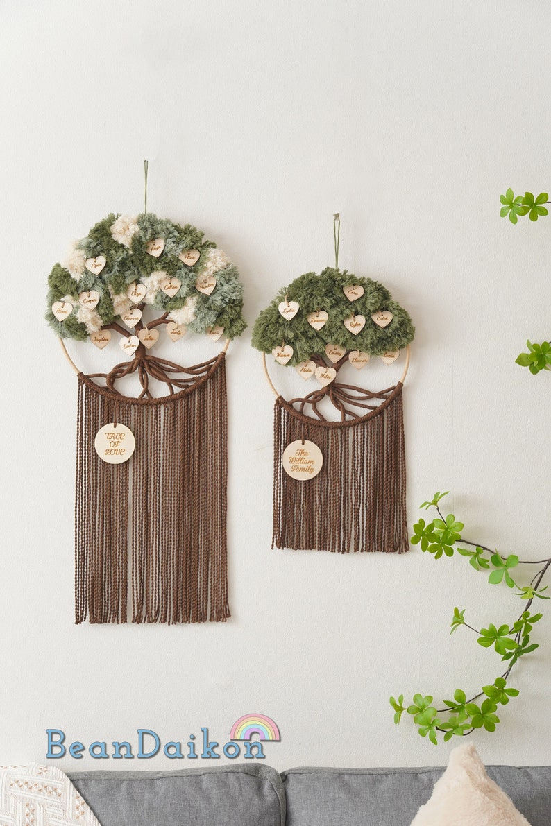 May include: Two green and white yarn pom-pom tree wall hangings with wooden hearts with names on them. The larger tree has the text "Tree of Love" on a wooden circle and the smaller tree has the text "The William Family" on a wooden circle.