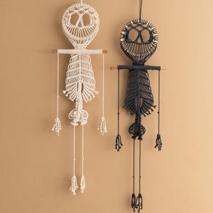 May include: Two macrame wall hangings, one white and one black, each featuring a skull design with a peace sign in the center. Both hangings have tassels and wooden dowels.