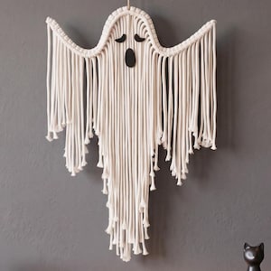 May include: A wall hanging crafted from off-white macrame cord, shaped like a ghost with outstretched arms. It features two black felt eyes and a black felt oval mouth. The design consists of knotted cord sections and long, loose hanging fringe.