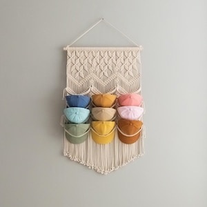 May include: A macrame wall hanging with a geometric pattern, featuring nine different colored baseball caps hanging from the bottom. The caps are in shades of blue, green, yellow, orange, and pink.