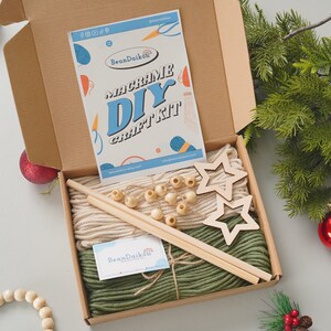 May include: A macrame DIY craft kit in a brown box with green and beige yarn, wooden dowels, wooden beads, and two wooden star shapes. The box is open and the contents are visible. The kit is from BeanDaikon.