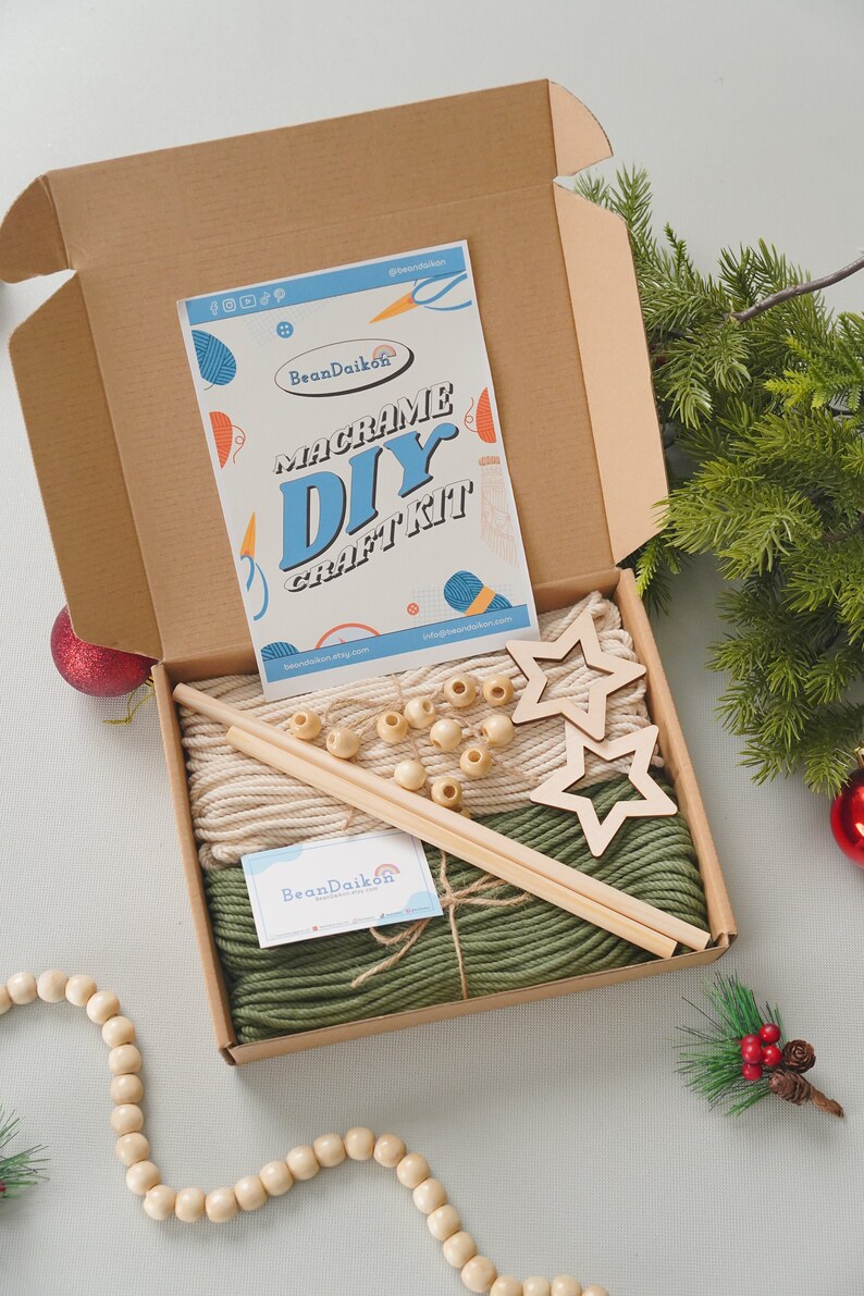 May include: A macrame DIY craft kit in a brown box with green and beige yarn, wooden dowels, wooden beads, and two wooden star shapes. The box is open and the contents are visible. The kit is from BeanDaikon.