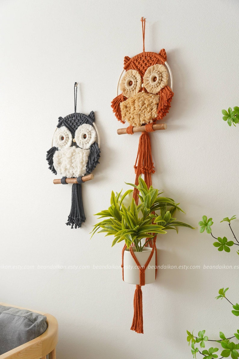 Peut inclure: Deux tentures murales en macram&eacute; en forme de hibou avec des chevilles en bois et des glands. Le hibou de gauche est gris et blanc, et le hibou de droite est orange et blanc. Une plante est suspendue au hibou orange.
