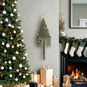 May include: A wall-hanging macrame Christmas tree in olive green cord, featuring a wooden star at the top, small wooden beads along the branches, and a circular wooden ornament with the text Merry Christmas hanging from the bottom.