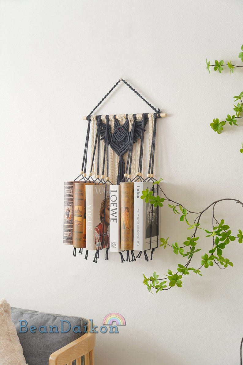 May include: A macrame wall hanging with a gray and white design holds a row of books. The books have titles like "Vogue", "LOEWE", and "Union" on their spines.