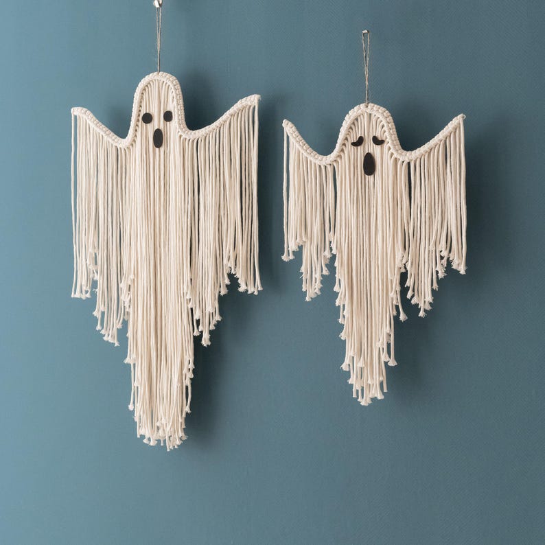 May include: Two white macrame ghost wall hangings with black eyes. The ghosts are made of yarn and have a fringe design.