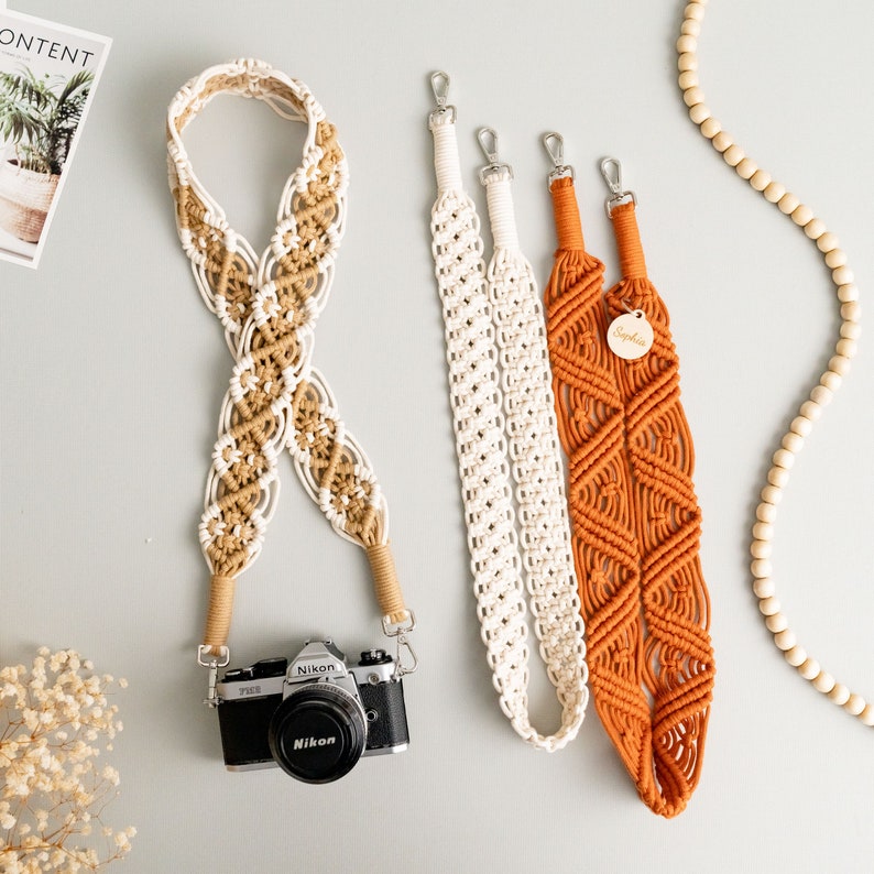 May include: Three camera straps made of macrame with a white and brown strap, a white strap, and an orange strap. The straps are hanging on a light gray background.