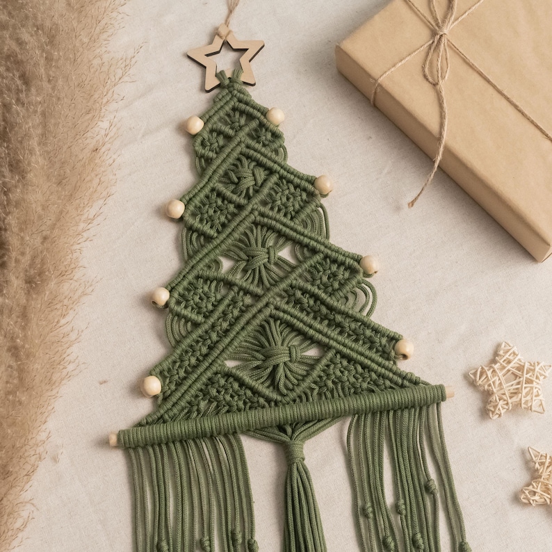 May include: A wall hanging shaped like a Christmas tree made of olive green macrame cord. The design features a wooden star at the top, light-coloured wooden beads along the edges, and a fringe at the bottom. The structure is composed of intricate knotted patterns.