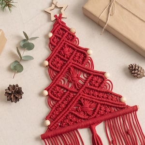 May include: A red macrame wall hanging shaped like a Christmas tree, featuring intricate knotted patterns, light-coloured wooden beads along the edges, a wooden star at the top, and a long fringe at the bottom suspended by a wooden dowel.
