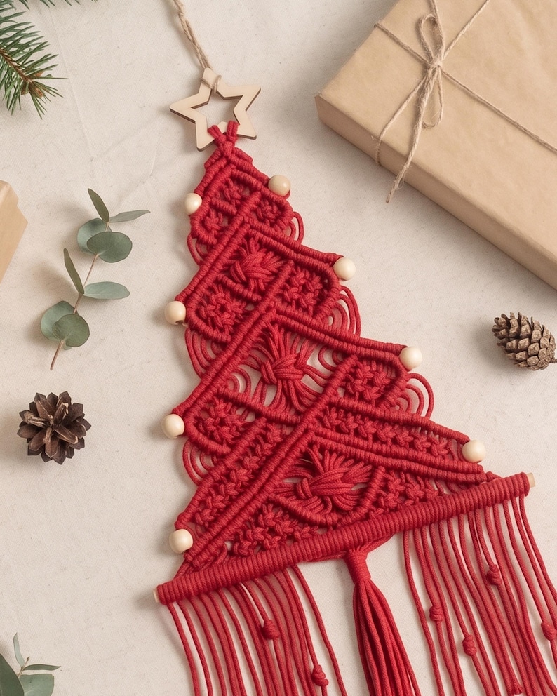 May include: A red macrame wall hanging shaped like a Christmas tree, featuring intricate knotted patterns, light-coloured wooden beads along the edges, a wooden star at the top, and a long fringe at the bottom suspended by a wooden dowel.