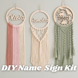 Puede incluir: Tres kits de letreros de nombre de bricolaje con aros de madera y flecos de macramé. Los aros están decorados con los nombres "Olivia", "Amelia" y "Robert". El aro "Robert" tiene un diseño de dinosaurio.