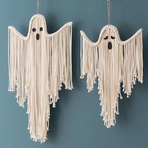 May include: Two white macrame ghost wall hangings with black eyes and mouths. The ghosts are made of yarn and have a fringe design.