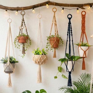 May include: Six macrame plant hangers in various colors including cream, tan, brown, and navy blue, suspended from a horizontal wooden branch with string lights. Each hanger holds a potted plant and features intricate knotted designs and tassels.