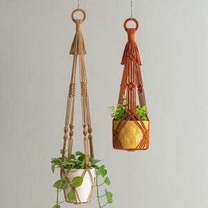 May include: Two macrame plant hangers, one in a natural colour and one in a burnt orange colour, hanging from the ceiling with plants in white and yellow pots.