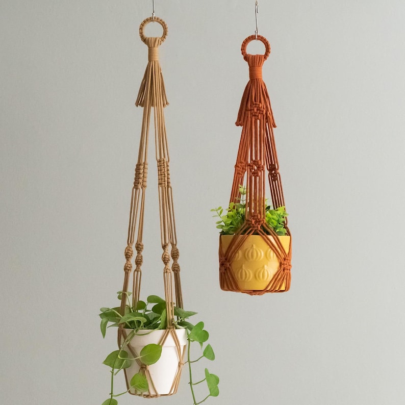 May include: Two macrame plant hangers, one in a natural color and one in a burnt orange color, hanging from the ceiling with plants in white and yellow pots.