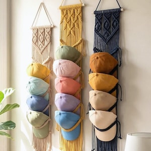 May include: Three macrame hat organisers in beige, mustard yellow, and navy blue, each holding several baseball caps in various colours. The caps include yellow, green, pink, orange, purple, and blue. The organisers hang on a white wall.