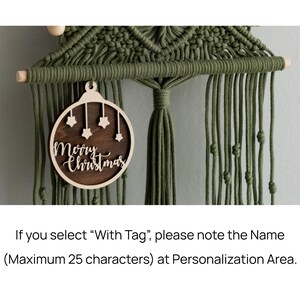 May include: A dark olive green macrame wall hanging featuring a wooden dowel and a circular wooden ornament. The ornament has a dark brown centre with light wood cutouts of four small stars and the text Merry Christmas.