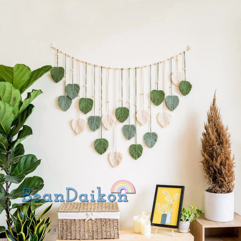 K&ouml;nnte beinhalten: Ein Wandbehang aus Makramee mit gr&uuml;nen und cremefarbenen Blattformen. Die Bl&auml;tter sind an einer wei&szlig;en Schnur mit Holzperlen befestigt. Der Wandbehang h&auml;ngt an einer wei&szlig;en Wand mit einer Pflanze und einem kleinen Holztisch mit einem Bilderrahmen und einem Weidenkorb.