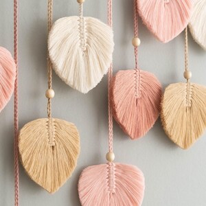 K&ouml;nnte beinhalten: Ein Wandbehang mit mehreren Makramee-Federformen in Rosa, Beige und Wei&szlig;. Die Federn sind an Schn&uuml;ren mit Holzperlen befestigt.