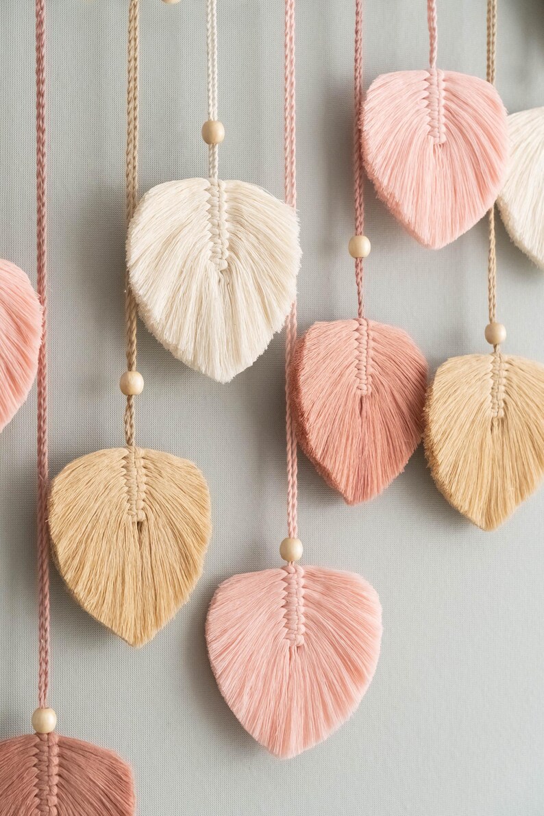 K&ouml;nnte beinhalten: Ein Wandbehang mit mehreren Makramee-Federformen in Rosa, Beige und Wei&szlig;. Die Federn sind an Schn&uuml;ren mit Holzperlen befestigt.