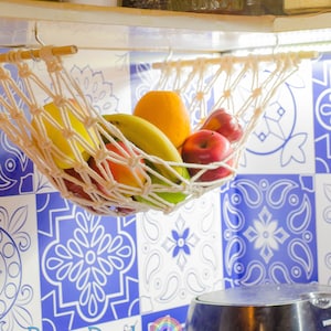 May include: A white macrame hammock hanging from a wooden shelf, filled with fresh fruit, including bananas, oranges, and apples. The hammock is hanging in front of a blue and white tiled wall.