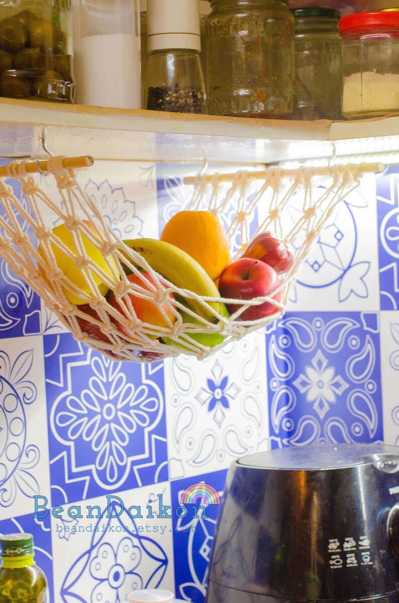 May include: A white macrame hammock hanging from a wooden shelf, filled with fresh fruit, including bananas, oranges, and apples. The hammock is hanging in front of a blue and white tiled wall.