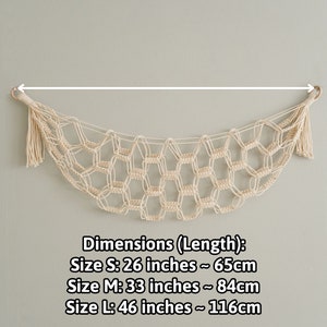 Op de afbeelding: Een beige macram&eacute; wanddecoratie met een kwast aan elk uiteinde. De decoratie heeft de vorm van een halve maan. Afmetingen (Lengte): Maat S: 65 cm Maat M: 84 cm Maat L: 116 cm
