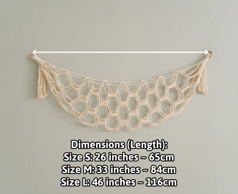 Op de afbeelding: Een beige macram&eacute; wanddecoratie met een kwast aan elk uiteinde. De decoratie heeft de vorm van een halve maan. Afmetingen (Lengte): Maat S: 65 cm Maat M: 84 cm Maat L: 116 cm