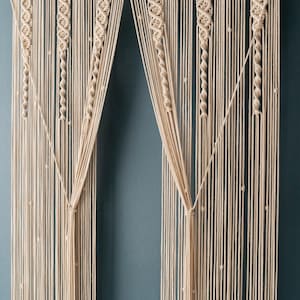 K&ouml;nnte beinhalten: Ein Makramee-Wandbehang mit geometrischem Muster. Der Behang ist aus naturfarbenem Baumwollschnur gefertigt und weist eine Reihe von Knoten und Schlaufen auf. Der Behang ist in zwei Abschnitte unterteilt, die jeweils ein anderes Muster aufweisen. Der Behang ist an einer Holzleiste aufgeh&auml;ngt.