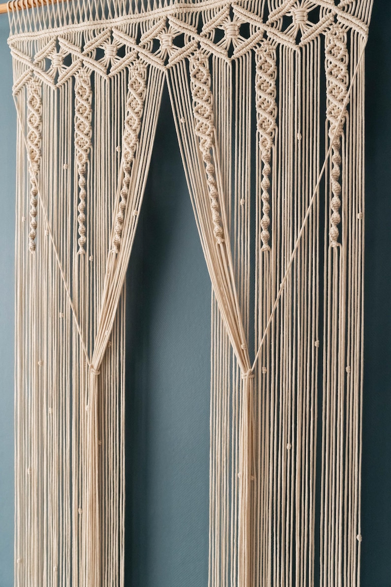 K&ouml;nnte beinhalten: Ein Makramee-Wandbehang mit geometrischem Muster. Der Behang ist aus naturfarbenem Baumwollschnur gefertigt und weist eine Reihe von Knoten und Schlaufen auf. Der Behang ist in zwei Abschnitte unterteilt, die jeweils ein anderes Muster aufweisen. Der Behang ist an einer Holzleiste aufgeh&auml;ngt.