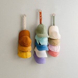 May include: Three wall-mounted hat organisers, each holding several baseball caps in shades of yellow, orange, green, blue, and pink. The organisers have decorative macrame or woven straps, and are designed to hold the caps neatly.