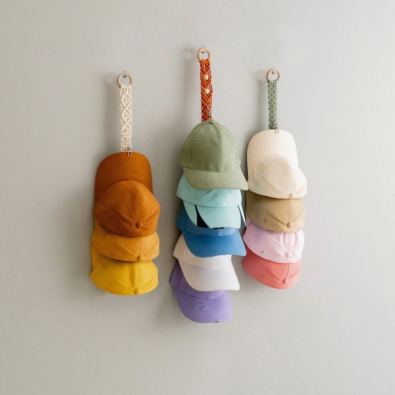 May include: Three wall-mounted hat organisers, each holding several baseball caps in shades of yellow, orange, green, blue, and pink. The organisers have decorative macrame or woven straps, and are designed to hold the caps neatly.