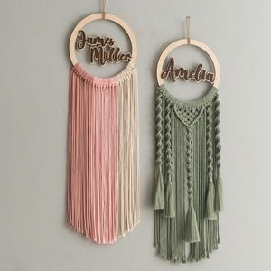 May include: Two macrame wall hangings with wooden hoops and personalized names. The left hanging has a pink and cream color scheme with the name "James Miller" on the hoop. The right hanging has a green color scheme with the name "Amelia" on the hoop.