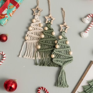 May include: Three macrame Christmas tree ornaments featuring knotted cotton cord in cream and two shades of green. Each tree is topped with a wooden star cutout and decorated with small wooden beads, finished with a hanging loop at the top.