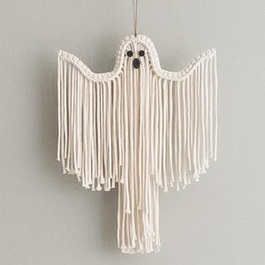 May include: White macrame ghost wall hanging with black eyes and a mouth.