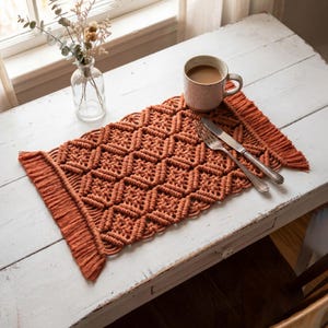 May include: A rectangular macrame placemat in a burnt orange color featuring a repeating diamond knot pattern and fringed edges. The placemat is displayed with a ceramic mug filled with coffee, a silver fork, and a silver knife placed on top.