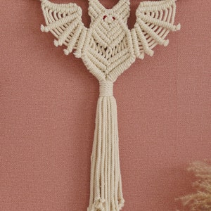 May include: A macrame bat wall hanging with a wooden dowel and a long tassel. The bat is made with natural coloured string and has red eyes.