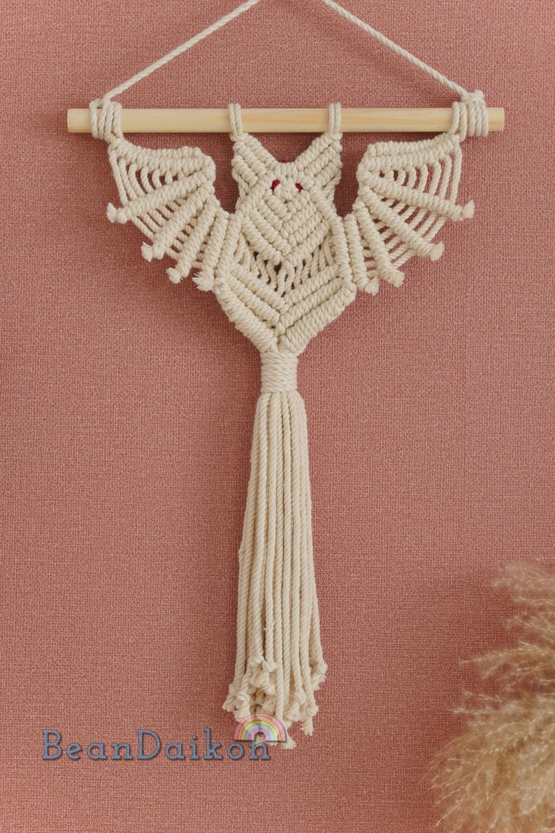 May include: A macrame bat wall hanging with a wooden dowel and a long tassel. The bat is made with natural coloured string and has red eyes.