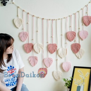 K&ouml;nnte beinhalten: Ein Wandbehang aus rosa und wei&szlig;em Garn mit einer Schnur aus Holzperlen. Der Behang hat die Form einer Reihe von Herzen, wobei jedes Herz einen Fransen aus Garn hat.