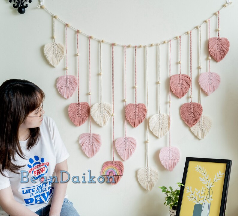K&ouml;nnte beinhalten: Ein Wandbehang aus rosa und wei&szlig;em Garn mit einer Schnur aus Holzperlen. Der Behang hat die Form einer Reihe von Herzen, wobei jedes Herz einen Fransen aus Garn hat.