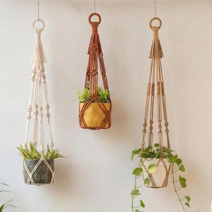May include: Three macrame plant hangers in cream, brown, and tan, each holding a potted plant. The hangers are suspended from the ceiling, showcasing a bohemian style. The pots are in shades of green, yellow, and white.