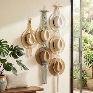 May include: A display of hats hanging on a wall. The hats are various colors, including beige and white, and are held by macrame hangers in shades of beige, green, and gold. The hats are displayed near a window with a view of a garden.