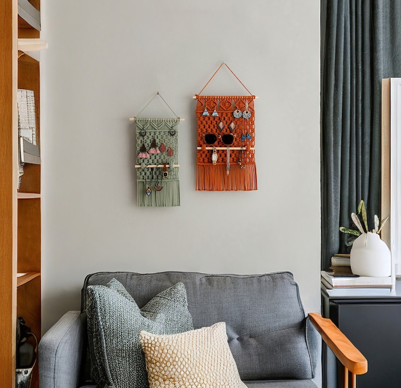 May include: Two macrame wall organisers with wooden dowels, one in green and one in orange, hanging on a white wall. The organisers have multiple compartments for storing jewellery, sunglasses, and other small items.