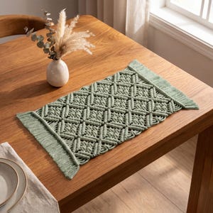 May include: A rectangular sage green macrame table runner featuring a repeating diamond knot pattern and fringed edges on both ends. The runner is crafted from thick cord and displays a textured, woven design that spans the length of the piece.