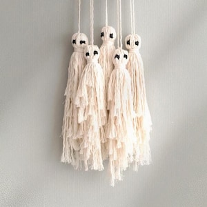 May include: A wall hanging featuring five off-white macrame ghost figures suspended by individual cords. Each ghost is crafted from knotted cotton cord with a rounded head and a long, fringed body, featuring two small black embroidered eyes.